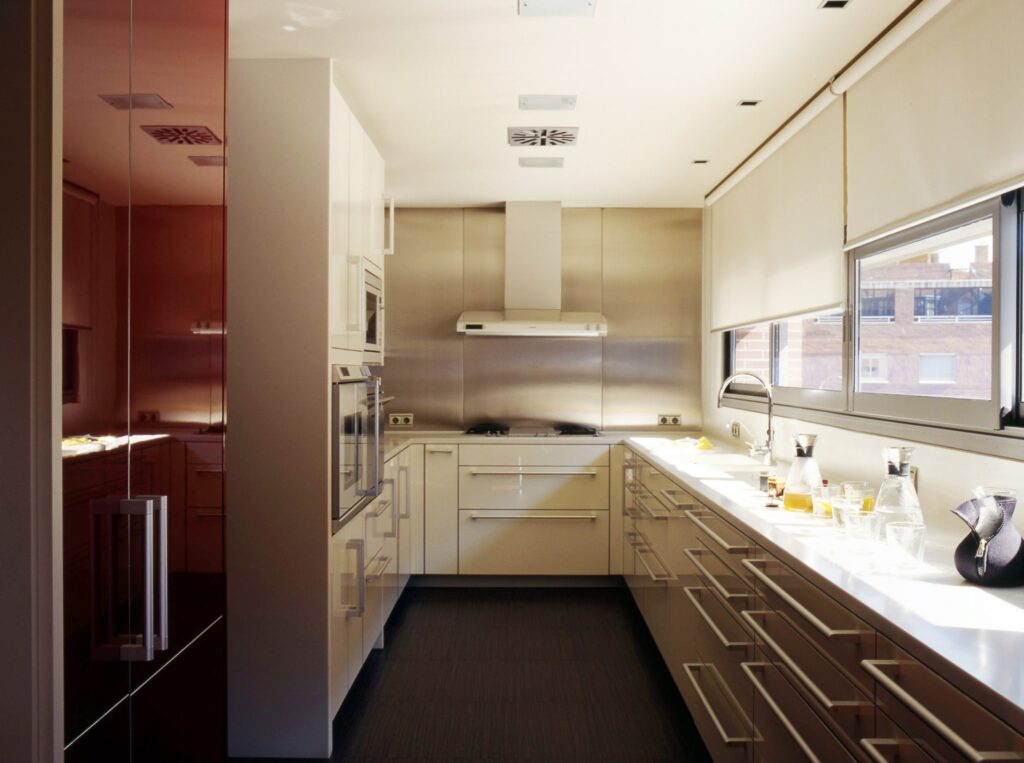 Cocina Minimalista Contemporánea con Elegantes Armarios Rojos y de Colores Claros Una cocina moderna que muestra un diseño minimalista con armarios reflectantes de color rojo profundo a la izquierda y armarios de colores claros con una encimera de acero inoxidable a la derecha. Una campana extractora está instalada sobre la estufa, y grandes ventanas permiten que la luz natural ilumine el espacio organizado adornado con artículos de cocina como recipientes de vidrio y utensilios.