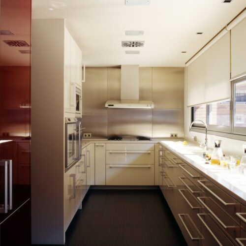 Una cocina moderna que muestra un diseño minimalista con armarios reflectantes de color rojo profundo a la izquierda y armarios de colores claros con una encimera de acero inoxidable a la derecha. Una campana extractora está instalada sobre la estufa, y grandes ventanas permiten que la luz natural ilumine el espacio organizado adornado con artículos de cocina como recipientes de vidrio y utensilios.