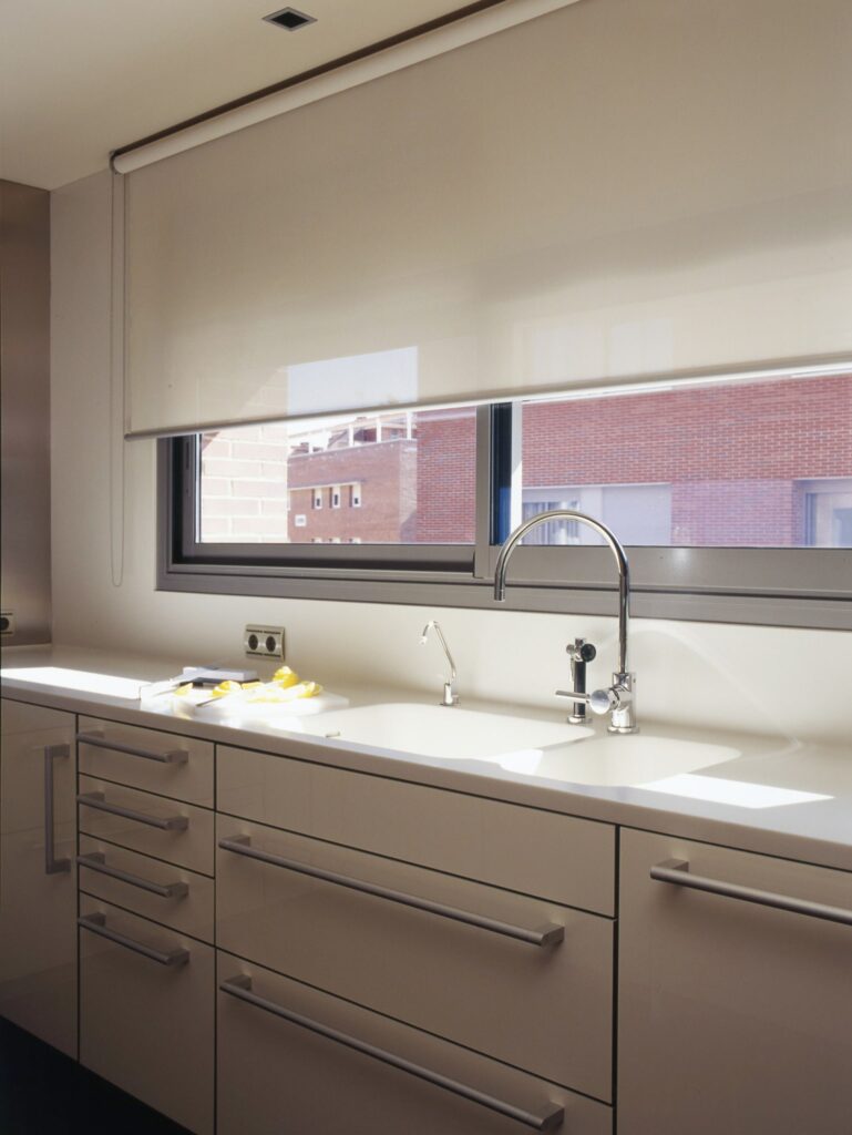 Cocina Moderna Elegante con Encimera Elegante y Luz Natural Un interior de cocina moderna que muestra una encimera elegante con un fregadero doble y un grifo de cromo pulido, rodeada de armarios minimalistas. La luz natural filtra a través de una gran ventana adornada con una persiana enrollable beige, proyectando un cálido resplandor sobre el espacio de trabajo. Elementos amarillos coloridos al lado del fregadero añaden un toque vibrante al diseño de cocina neutral que enfatiza la limpieza y la estética contemporánea.