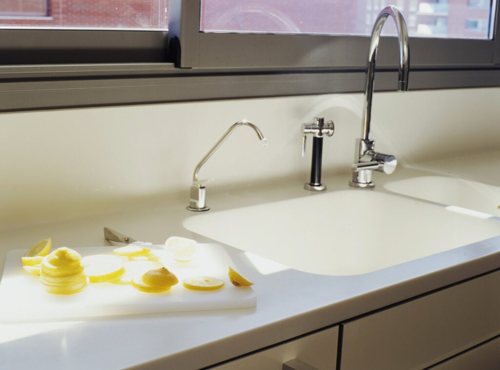 Fregadero de cocina moderno y elegante con grifos duales y limones frescos Un área de fregadero de cocina contemporáneo que muestra un diseño estilizado con una encimera blanca, grifos de cromo pulido y negros. Una tabla de cortar sostiene limones en rodajas y espirales, iluminados por la luz natural, destacando la estética minimalista.
