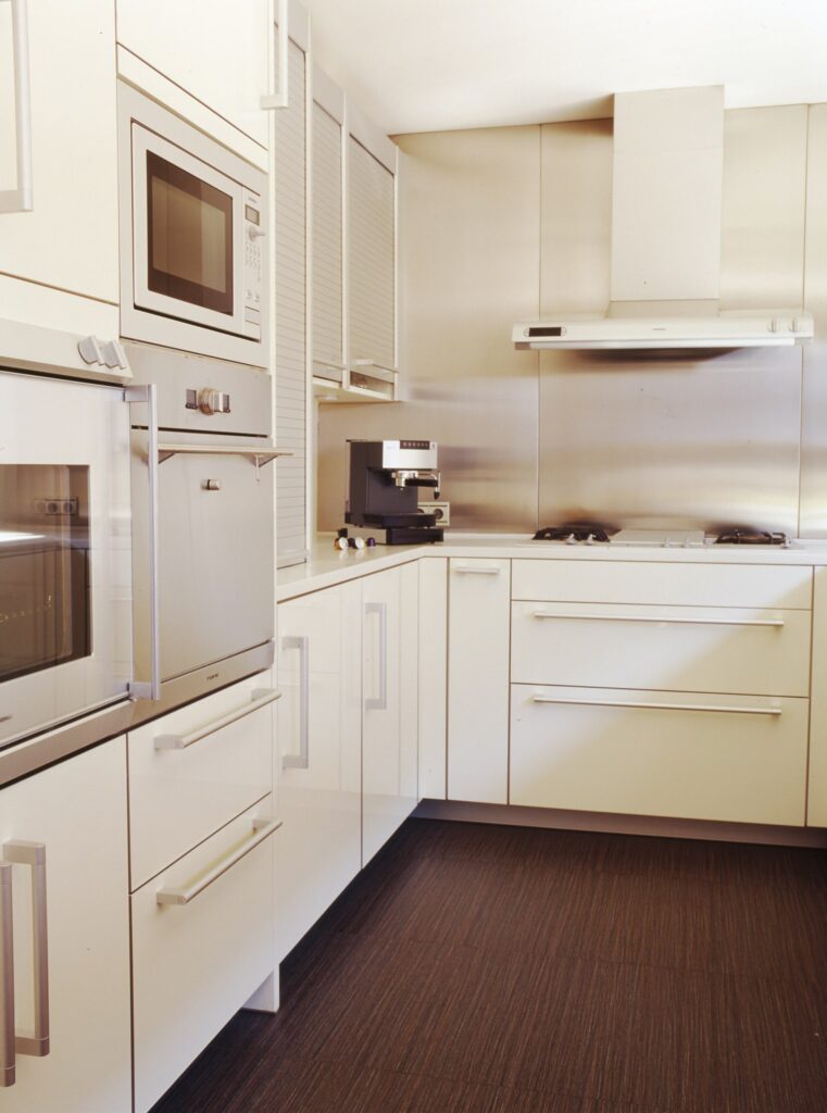 Diseño de cocina moderna con elegantes gabinetes blancos y electrodomésticos de acero inoxidable Cocina contemporánea que presenta gabinetes blancos, microondas de acero inoxidable, horno y máquina de café, combinados con un piso de madera oscura para un contraste elegante.