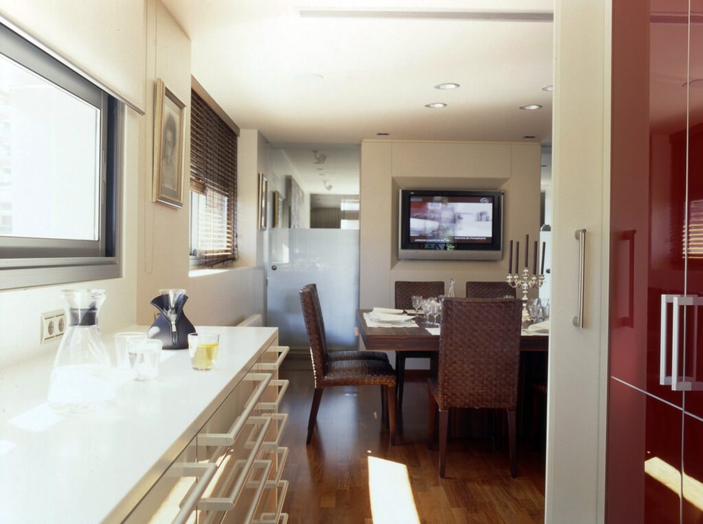 Área de Comedor Moderno y Minimalista con Luz Natural Una elegante área de comedor minimalista que presenta una encimera blanca con cristalería, incluida una jarra y pequeños vasos. En el fondo, una mesa de comedor de madera está dispuesta con utensilios elegantes y rodeada de sillas de mimbre. Un televisor de pantalla plana está montado en la pared, iluminado por la luz natural que entra a través de grandes ventanas que realzan la estética contemporánea del espacio.