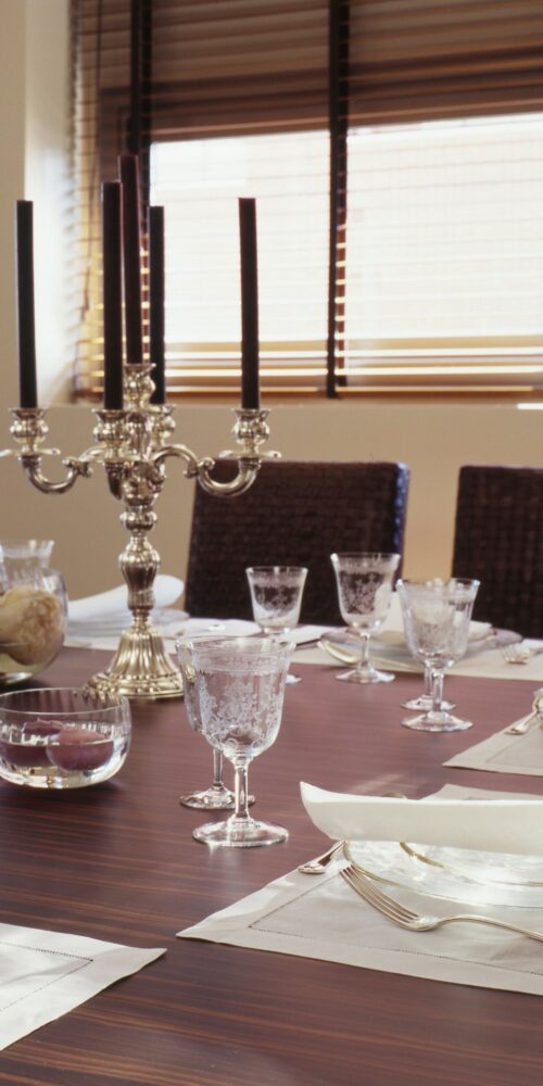 Una sofisticada mesa de comedor dispuesta con un elegante candelabro de plata que sostiene velas negras, adornada con cristalería ornamentada, copas de cristal y un simple camino de mesa blanco, iluminada por la cálida luz del sol que se filtra a través de persianas de madera.