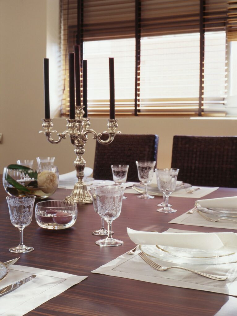 Configuraión Elegante de Mesa de Comedor con Candelabro de Plata y Cristalería de Cristal Una sofisticada mesa de comedor dispuesta con un elegante candelabro de plata que sostiene velas negras, adornada con cristalería ornamentada, copas de cristal y un simple camino de mesa blanco, iluminada por la cálida luz del sol que se filtra a través de persianas de madera.