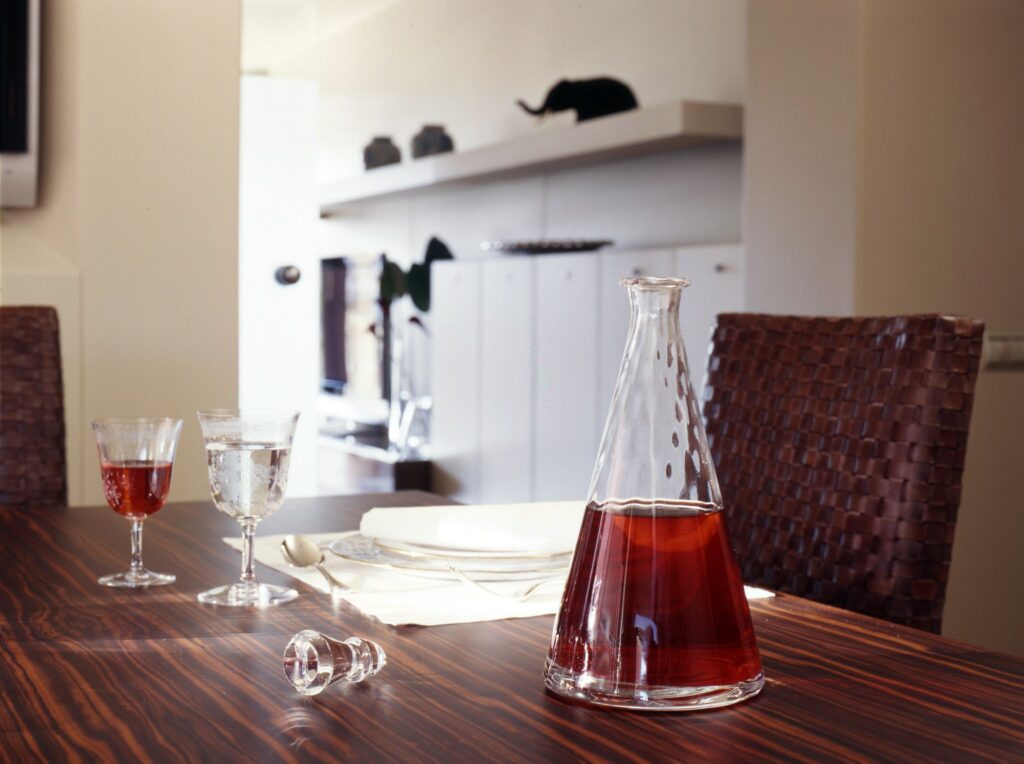 Configuración Elegante de Mesa de Comedor con Jarra de Vidrio y Fondo de Cocina Moderna Una mesa de comedor elegante que presenta una jarra de vidrio transparente llena de una bebida roja profunda, acompañada de dos copas: una con bebida roja y la otra con agua, sobre una superficie de madera rica y una cocina moderna con armarios blancos.
