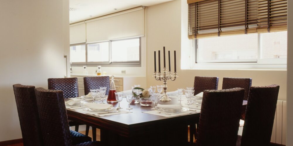 Comedor moderno que presenta una mesa de comedor de madera oscura dispuesta para una comida, rodeada de ocho sillas tejidas. La mesa está elegantemente decorada con platos blancos, cristalería brillante y un sofisticado candelabro de plata con velas negras. La luz natural se filtra a través de grandes ventanas con persianas de madera, iluminando la atmósfera acogedora del espacio.