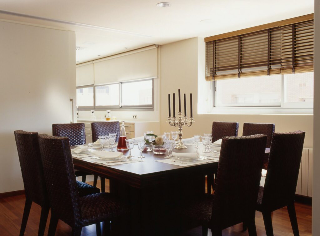 Comedor Moderno Elegante con Mesa de Madera Oscura y Sillas Tejidas Comedor moderno que presenta una mesa de comedor de madera oscura dispuesta para una comida, rodeada de ocho sillas tejidas. La mesa está elegantemente decorada con platos blancos, cristalería brillante y un sofisticado candelabro de plata con velas negras. La luz natural se filtra a través de grandes ventanas con persianas de madera, iluminando la atmósfera acogedora del espacio.