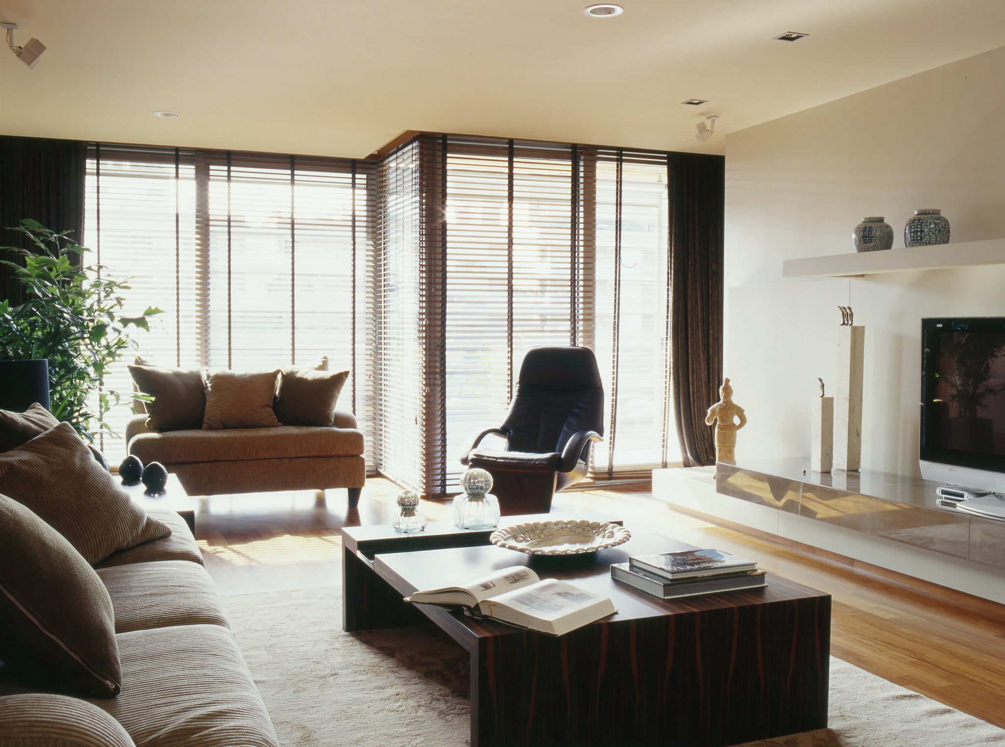 Una sala de estar contemporánea con grandes ventanales con persianas horizontales, un sofá cómodo con cojines decorativos, una silla de lounge negra elegante y una mesa de café moderna adornada con libros y elementos decorativos. El espacio cuenta con un cálido piso de madera, una suave alfombra y decoraciones minimalistas, incluyendo esculturas y jarrones sobre un televisor, creando una atmósfera acogedora e invitadora.