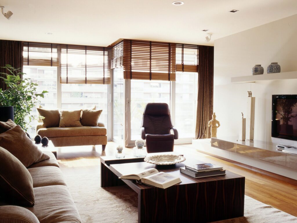 Sala de Estar Moderna y Estilosa con Luz Natural Una sala de estar moderna con una gran ventana con persianas de madera, un sofá acogedor con cojines mullidos, una mesa de café minimalista, una silla elegante, un cálido suelo de madera, una suave alfombra y jarrones decorativos y una escultura, además de plantas verdes.