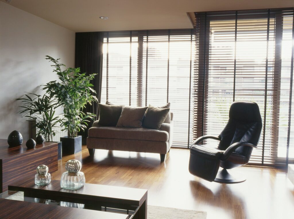 Salón moderno y elegante con sofá marrón acogedor y plantas verdes exuberantes Un salón contemporáneo con un sofá marrón con cojines grises, una silla reclinable negra y grandes ventanas con persianas de madera, que dejan entrar una suave luz natural. El cuarto incluye una mesa de café de madera con elementos decorativos y vibrantes plantas verdes, creando una atmósfera cálida y acogedora.