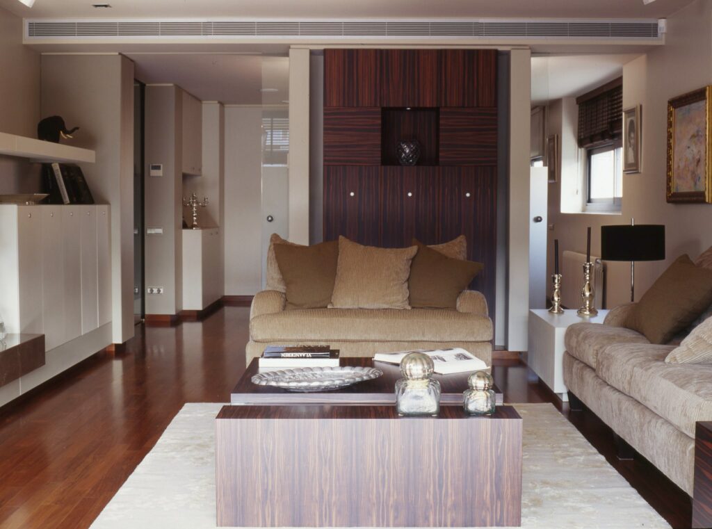 Sala de estar moderna y elegante con elementos de diseño contemporáneo Una sala de estar moderna y elegante que presenta una mesa de café de madera oscura sobre una suave alfombra, dos sofás de tela texturizada adornados con cojines decorativos y una pared con paneles de madera que cuenta con estanterías. La luz natural brillante inunda el espacio a través de grandes ventanas, resaltando los sofisticados elementos decorativos, incluidas jarrones y candelabros metálicos.