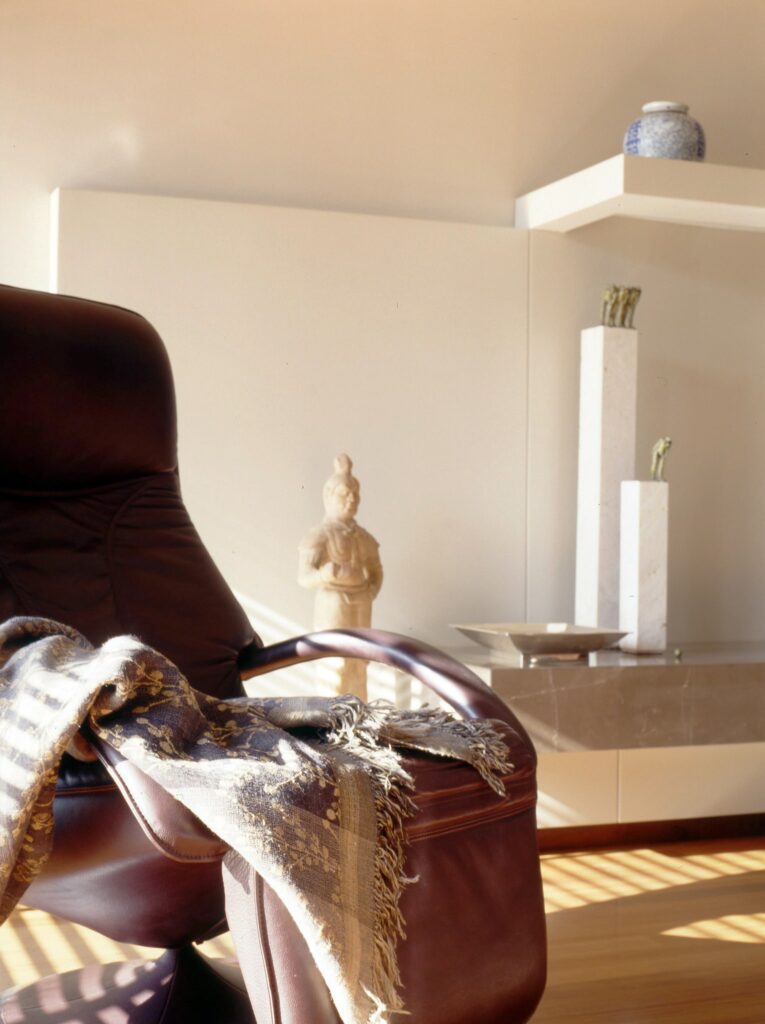 Espacio moderno y acogedor con sillón de felpa y chal decorativo Una sala de estar moderna con un sillón marrón de felpa cubierto con un chal decorativo, acompañado de una estantería minimalista que exhibe un jarrón de cerámica azul y una pequeña estatua, iluminada por luz natural suave.