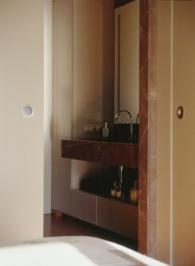 Baño Minimalista Moderno con Encimera de Mármol Oscuro Vista interior de un baño minimalista moderno que muestra una elegante encimera de mármol oscuro con un lavabo estilizado, iluminado por luz natural. El espacio presenta colores de pared neutros y una disposición cuidadosamente seleccionada de pequeñas botellas en la encimera.