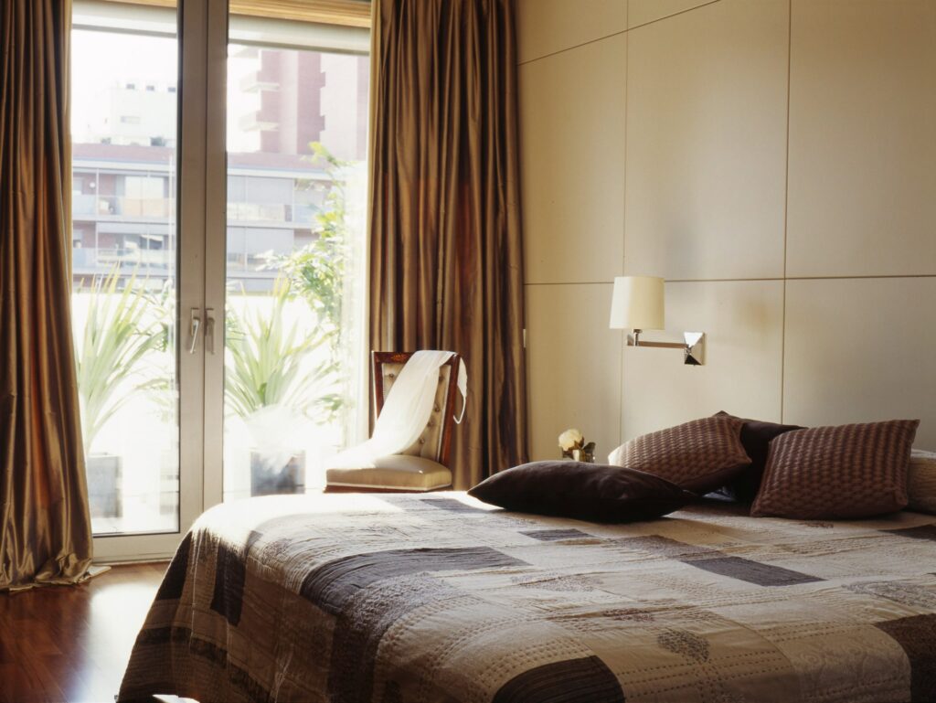 Dormitorio Moderno Elegante con Luz Natural y Decoración Elegante Un dormitorio contemporáneo que presenta una cama bien hecha con ropa de cama estampada y cojines decorativos, iluminado por la luz natural que entra a través de grandes puertas de vidrio. La habitación incluye elegantes cortinas drapeadas, una silla blanca estilosa, paredes de color claro y una vista del balcón adornada con plantas.