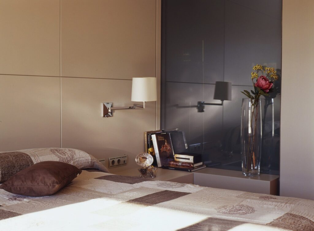 Dormitorio Moderno Acogedor con Colcha de Parchwork y Decoración Elegante Un dormitorio sereno con una cama bien hecha adornada con una colcha de parchwork y un cojín decorativo marrón, una lámpara de pared que proporciona luz suave, una mesa de noche con una pila de libros y un objeto decorativo, y un jarrón de vidrio alto con una flor rosa y vegetación, todo enmarcado por una pared en tonos neutros que brinda una atmósfera limpia y acogedora.