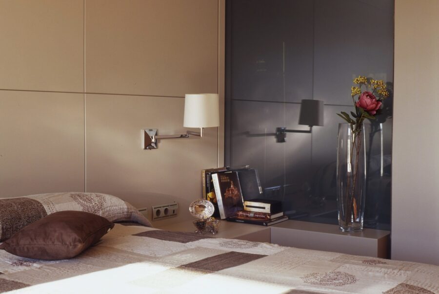 Un dormitorio sereno con una cama bien hecha adornada con una colcha de parchwork y un cojín decorativo marrón, una lámpara de pared que proporciona luz suave, una mesa de noche con una pila de libros y un objeto decorativo, y un jarrón de vidrio alto con una flor rosa y vegetación, todo enmarcado por una pared en tonos neutros que brinda una atmósfera limpia y acogedora.