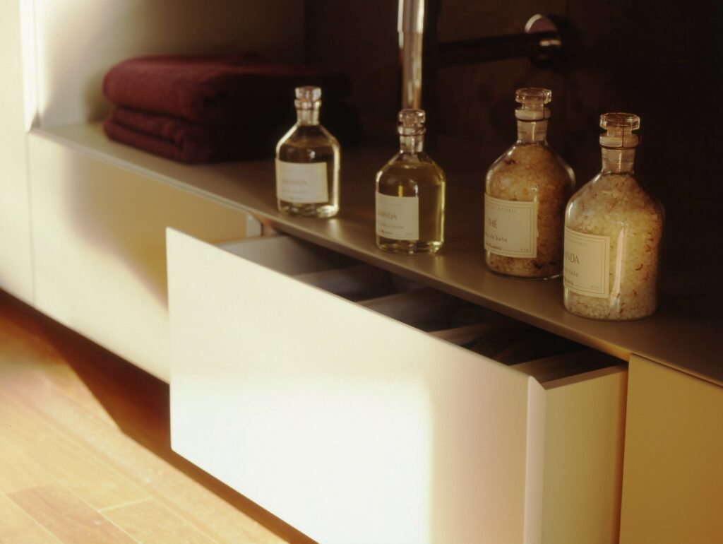 Interiores Modernos Elegantes con Armario Minimalista y Luz Suave Una escena interior moderna que presenta un elegante suelo de madera, un armario minimalista con un cajón blanco parcialmente abierto que muestra un interior ordenado, y toallas oscuras apiladas ordenadamente. Botellas de vidrio con etiquetas sencillas que contienen líquido y sales de baño están sobre el armario, todo iluminado por una luz suave que crea una atmósfera acogedora.