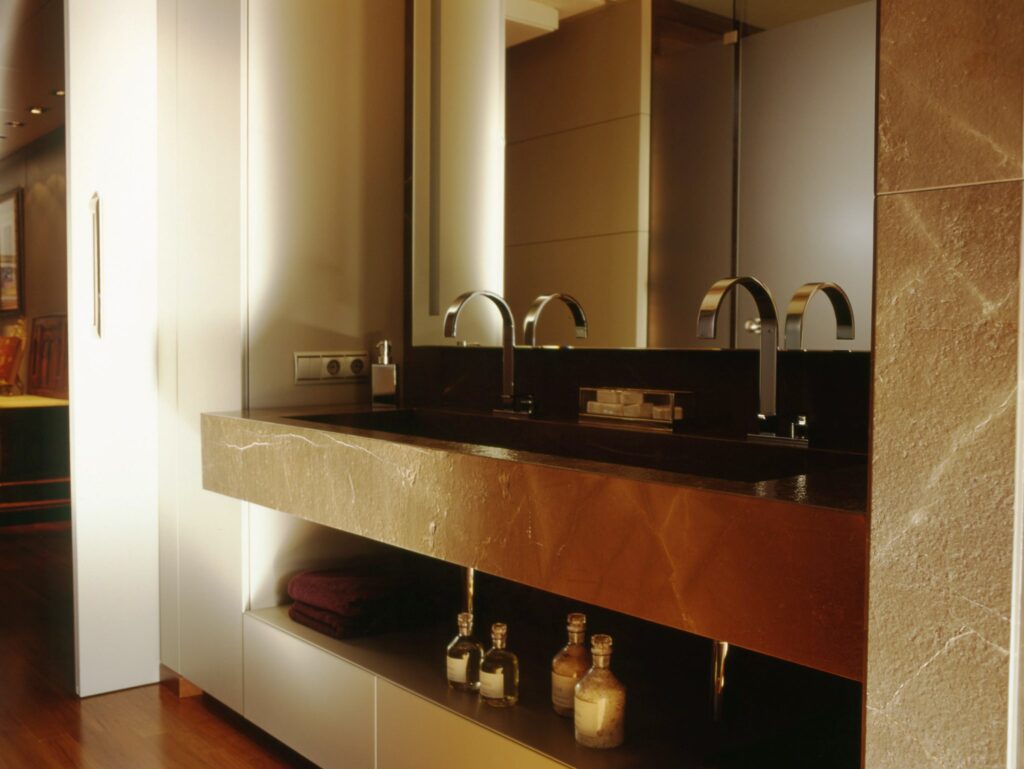 Fregadero de baño moderno y elegante con encimera de mármol oscuro y grifos de cromo Un área de fregadero de baño moderno que presenta una elegante encimera de mármol oscuro, dos grifos de cromo, botellas de vidrio y toallas enrolladas, todo enmarcado por un mueble claro y un gran espejo.
