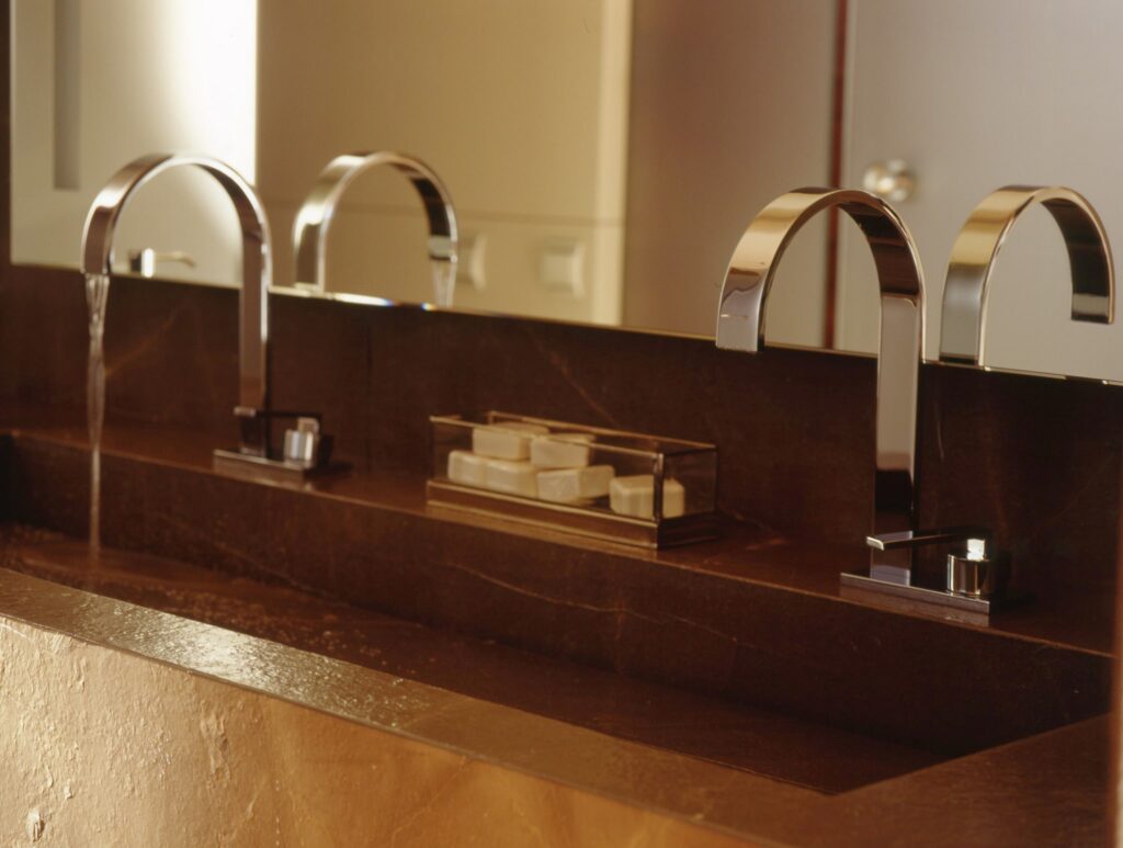 Baño moderno con grifos de cromo elegantes y lavabo de piedra oscura Baño contemporáneo que presenta un elegante lavabo moderno con grifos de cromo dobles y un lavabo de piedra oscura pulida, resaltado por una cálida iluminación ambiental de un espejo suavemente iluminado y jabones organizados de manera ordenada.