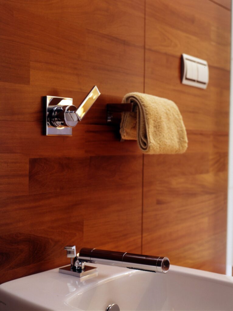 Baño contemporáneo con lavabo blanco elegante y pared de paneles de madera Un baño moderno y elegante que muestra un lavabo blanco con un grifo de manija de madera, una pared de paneles de madera de diseño elegante, y una toalla beige doblada con cuidado en una barra de toallas cromada, destacando el minimalismo y la estética contemporánea.