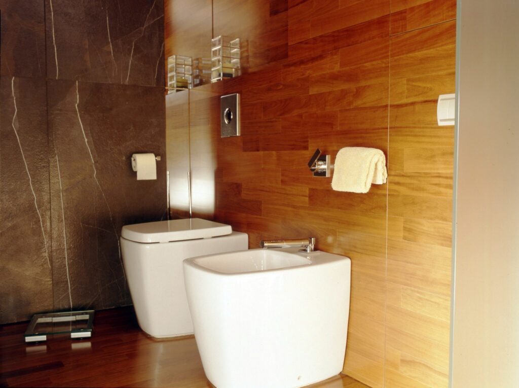 Diseño de Baño Minimalista Contemporáneo con Accesorios Blancos y Acabados de Madera Baño moderno con un diseño elegante y paredes texturizadas oscuras, paneles de madera, inodoro y bidé blancos, y un elegante toallero.
