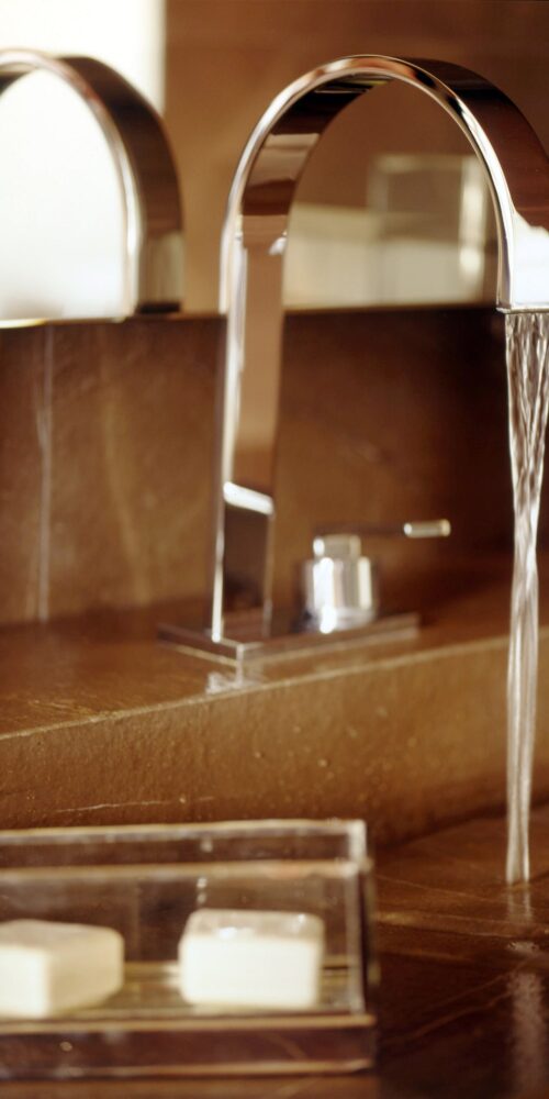 Un grifo cromado elegante con un pico curvado vierte agua en un lavabo de baño moderno, complementado por una encimera de piedra natural y dos pastillas de jabón rectangulares en una bandeja clara.