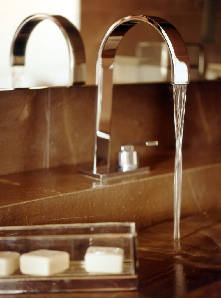 Lavabo moderno y elegante con grifo cromado y encimera de piedra natural Un grifo cromado elegante con un pico curvado vierte agua en un lavabo de baño moderno, complementado por una encimera de piedra natural y dos pastillas de jabón rectangulares en una bandeja clara.