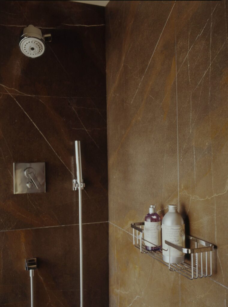 Espacio de Ducha Moderno y Lujoso con Paredes de Mármol Marrón Área de ducha contemporánea que muestra elegantes paredes de mármol marrón, un cabezal de ducha de lluvia montado en el techo, un grifo plateado elegante y una unidad de ducha de mano, con un estante de ducha de cromo que contiene botellas de champú y acondicionador minimalistas en púrpura y blanco.