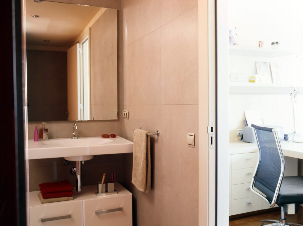 Baño Moderno Estiloso con Lavabo Minimalista y Espacio de Trabajo Un baño contemporáneo que presenta un lavabo y un espejo minimalistas, toallas rojas y beige organizadas ordenadamente en un gabinete, y un vistazo a un espacio de trabajo ordenado con un escritorio blanco, silla ergonómica y suministros de oficina, todo mostrando una atmósfera brillante y acogedora.