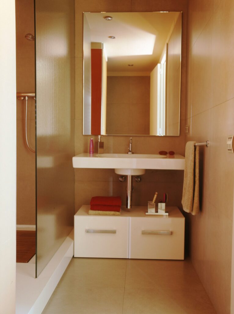 Baño Moderno Minimalista con Diseño Elegante Baño contemporáneo con un gran espejo rectangular, lavabo blanco suspendido y mampara de ducha de vidrio. El espacio incluye cajones de almacenamiento y elementos decorativos, todo en una brillante y suave paleta de colores beige y tonos claros.