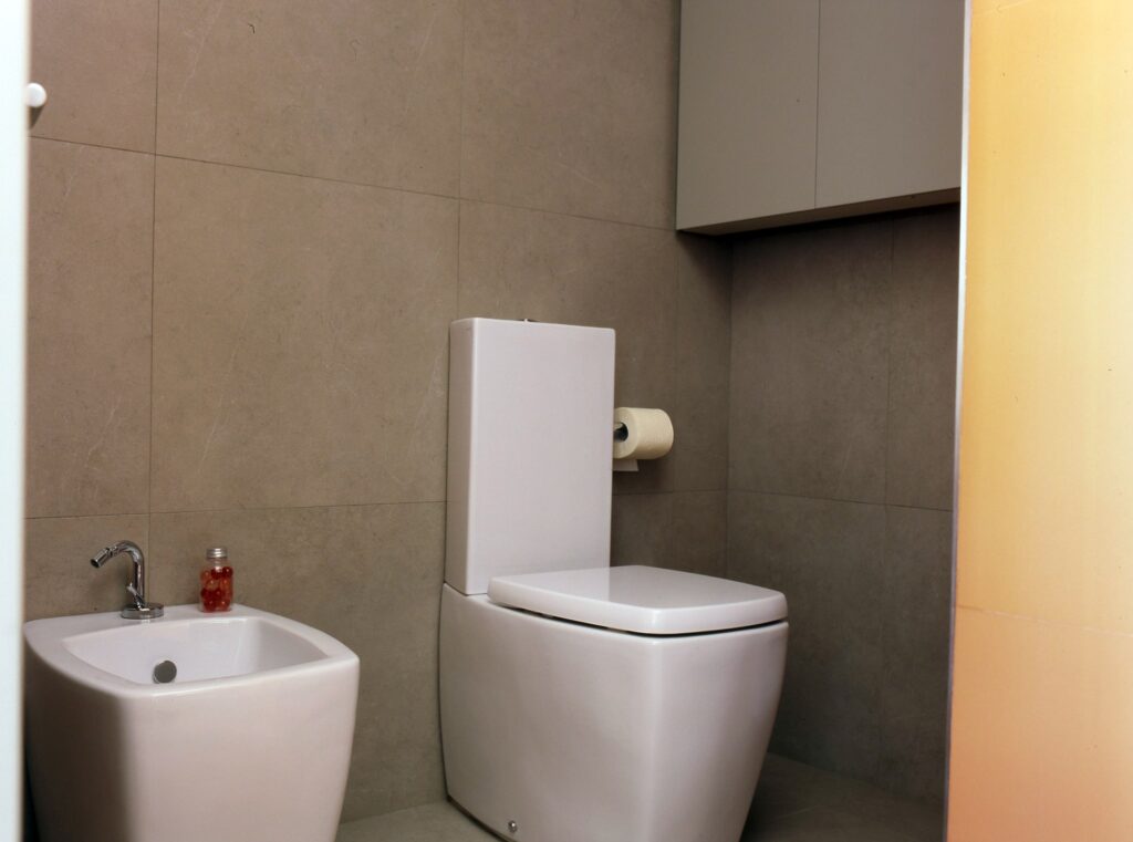 Baño Minimalista Moderno con Inodoro Blanco y Bidé Un baño brillante y moderno que presenta un elegante inodoro blanco y un estiloso bidé situado contra una pared de azulejos de tonos neutros. El espacio bien iluminado cuenta con un rollo de papel higiénico montado al lado del inodoro, y un pequeño recipiente de jabón líquido colocado de manera elegante sobre el bidé. Encima del inodoro, un armario contemporáneo mejora el almacenamiento mientras mantiene una estética limpia y minimalista.