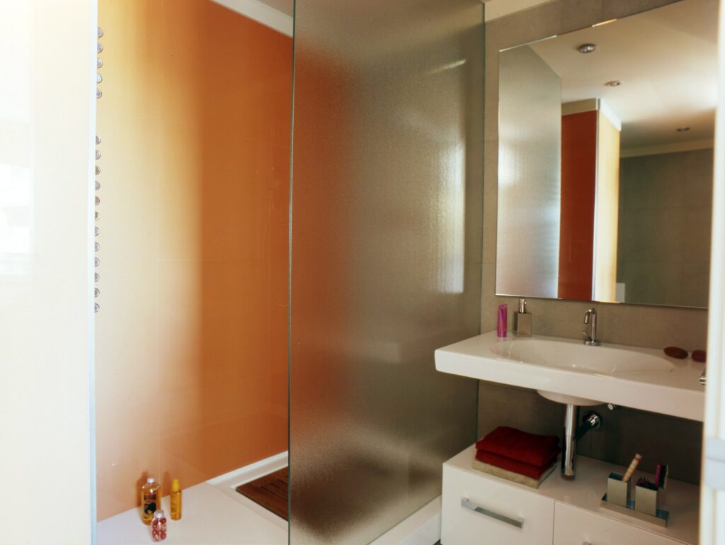 Diseño de baño moderno con ducha de cristal y lavabo elegante Baño moderno y espacioso que presenta un área de ducha con paredes de cristal a la izquierda y un lavabo rectangular elegante a la derecha, adornado con un gran espejo que refleja las cálidas paredes naranjas y un suelo de baldosas blancas, complementado por artículos de tocador organizados ordenadamente y toallas de colores.