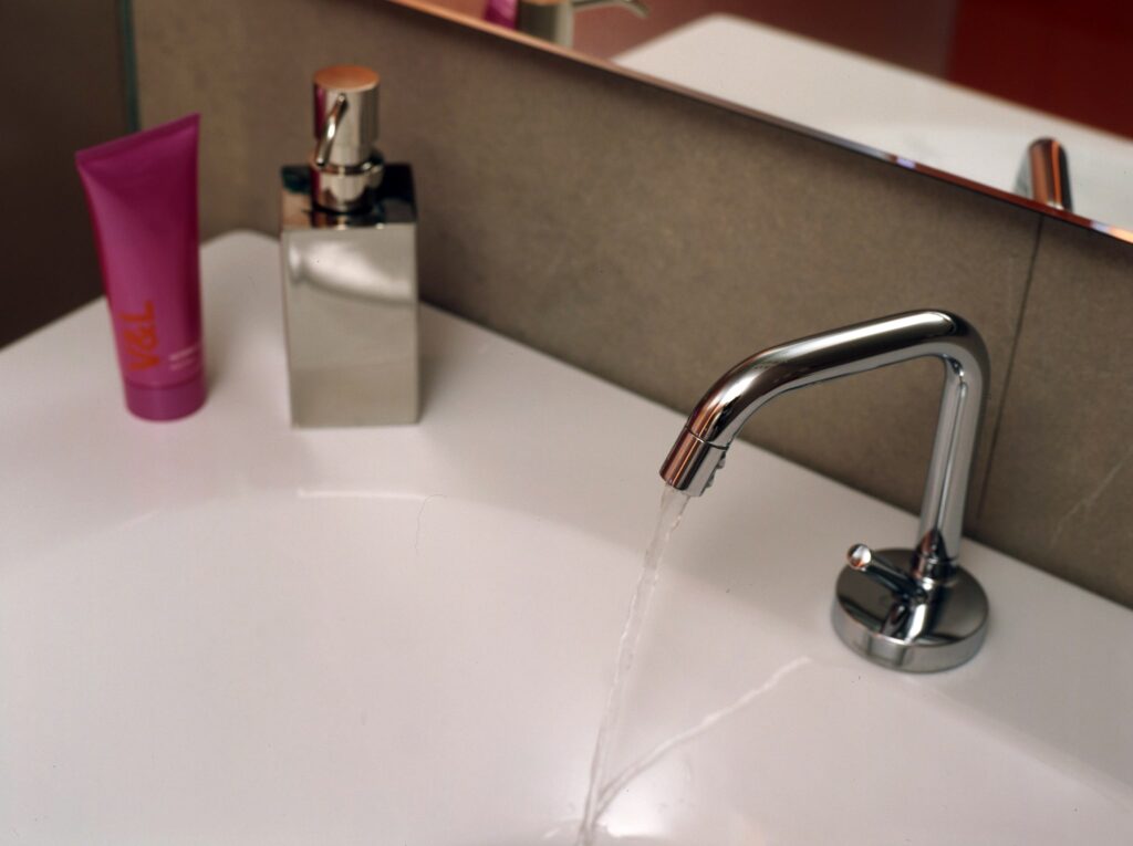 Lavabo moderno con grifo cromado y accesorios elegantes Un lavabo moderno y elegante que presenta un grifo cromado con agua fluyendo, un tubo de loción rosa y un dispensador de jabón plateado brillante sobre una encimera blanca lisa, con un espejo en el fondo que refleja el elegante baño.