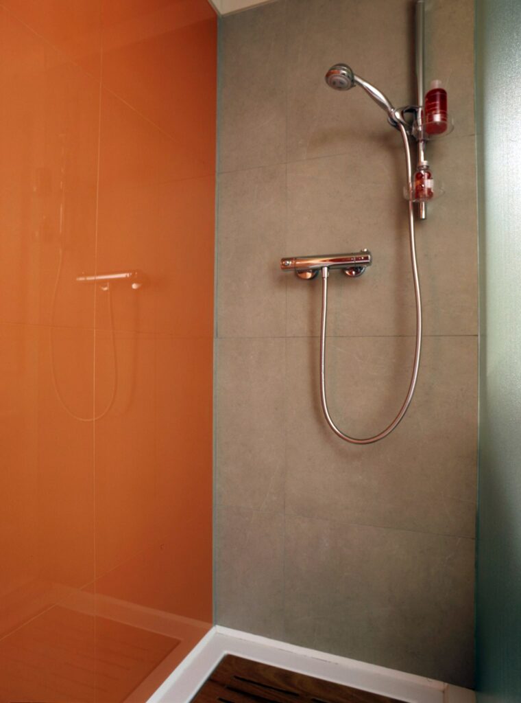Diseño de Ducha Moderna y Elegante con Pared de Vidrio Naranja y Pared Texturizada Gris Una ducha contemporánea con una impactante pared de vidrio naranja, que contrasta con una pared texturizada gris, una ducha con cabezal de cromo y un suelo de ducha con textura de madera, resaltando la estética minimalista.
