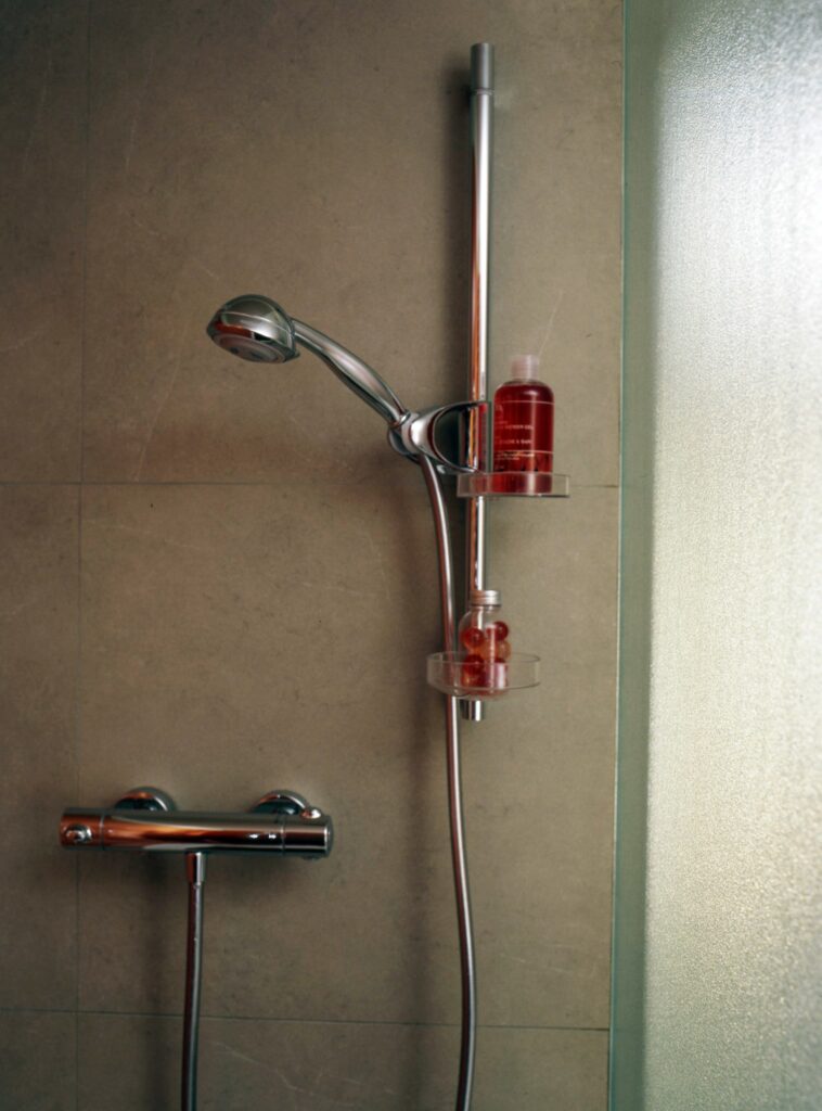 Configuración Moderna de Ducha de Baño con Accesorios de Cromo y Diseño Elegante Una configuración de ducha contemporánea que presenta una ducha de cromo pulido, un panel de control y una elegante estantería transparente que sostiene una botella roja, todo contra un fondo de azulejos neutros.