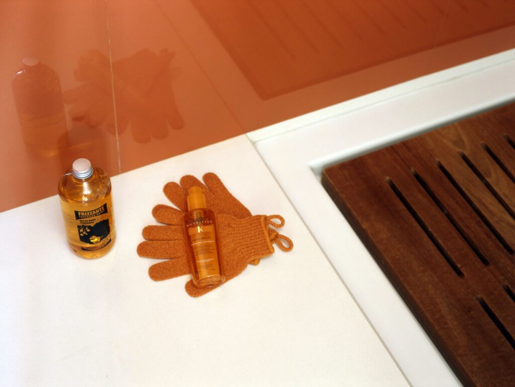 Frizzante y Productos para el Cuidado del Cabello con Guantes Exfoliantes sobre una Superficie Elegante Una botella transparente de Frizzante con un diseño negro y amarillo y una botella de producto para el cabello transparente colocada sobre una superficie blanca junto a suaves guantes exfoliantes naranjas, con un mural cálido de color naranja de fondo.