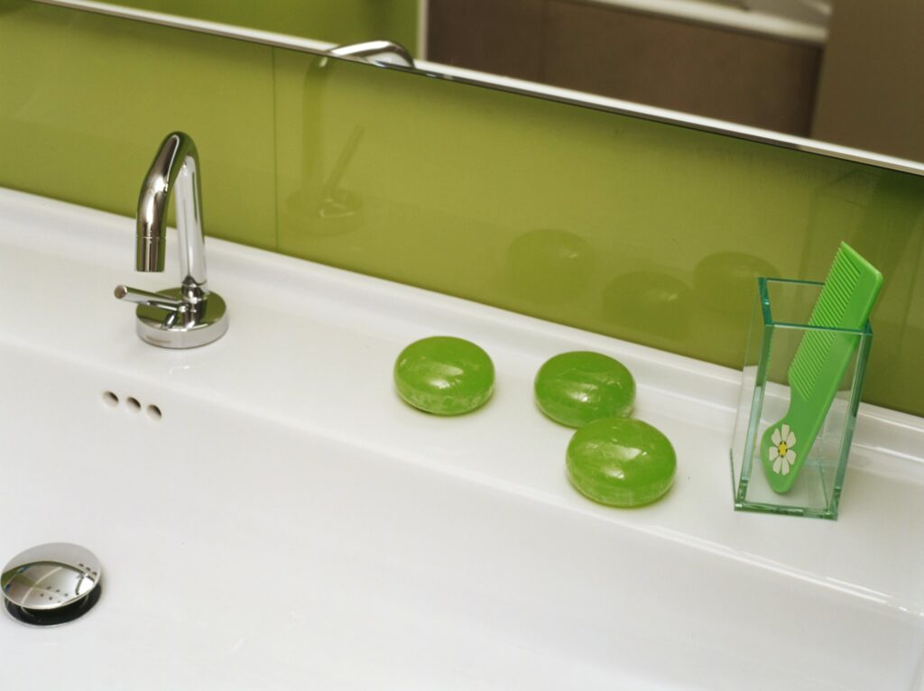 Fregadero de Baño Moderno y Elegante con Accesorios Verdes Fregadero de baño moderno con un grifo cromado, barras de jabón verde ordenadamente dispuestas sobre una encimera blanca, un peine verde con diseño floral en un recipiente transparente y un panel verde liso en el fondo para una estética fresca.