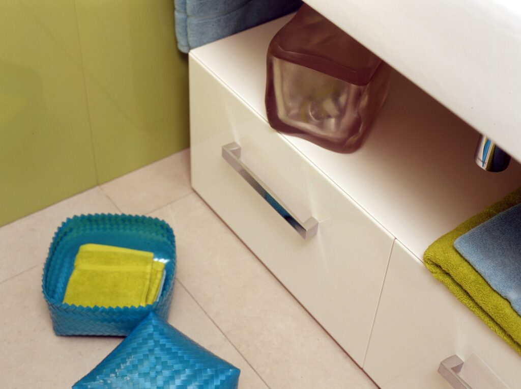 Baño Organizado con Almacenamiento Colorido y Vanity Elegante Un baño ordenado que muestra dos contenedores de almacenamiento coloridos, una cesta cuadrada texturizada azul con una toalla amarilla doblada, y un contenedor marrón debajo de un vanity blanco. El vanity presenta un diseño moderno con un mango plateado y está cubierto con toallas dobladas ordenadamente en verde y azul. El fondo destaca un suelo de azulejos neutros y una pared verde calmada.