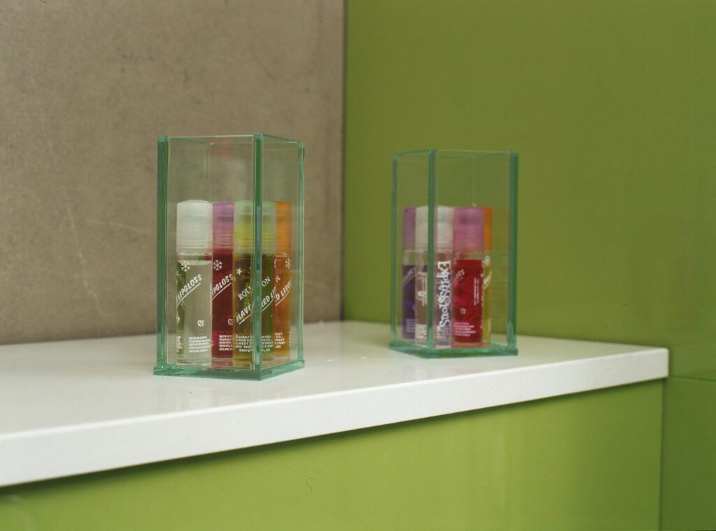 Botellas Coloridas de Productos de Belleza en Vitrinas Transparentes Una exhibición organizada de coloridas botellas de belleza y cuidado de la piel en dos vitrinas transparentes sobre una encimera blanca, con un suave fondo verde.