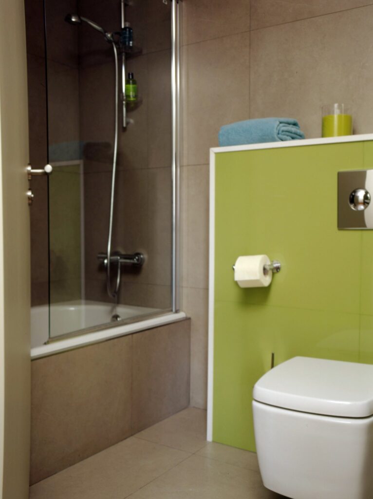 Diseño de Baño Moderno con Ducha de Cristal y Acentos Verdes Vibrantes Un baño contemporáneo que muestra un cabina de ducha de cristal equipada con una ducha de mano, junto a una bañera elegante. Un inodoro blanco está posicionado al lado de una vibrante pared verde que exhibe dos toallas azules ordenadamente apiladas y una botella decorativa. El suelo de baldosas de tonos neutros realza la estética limpia del espacio.