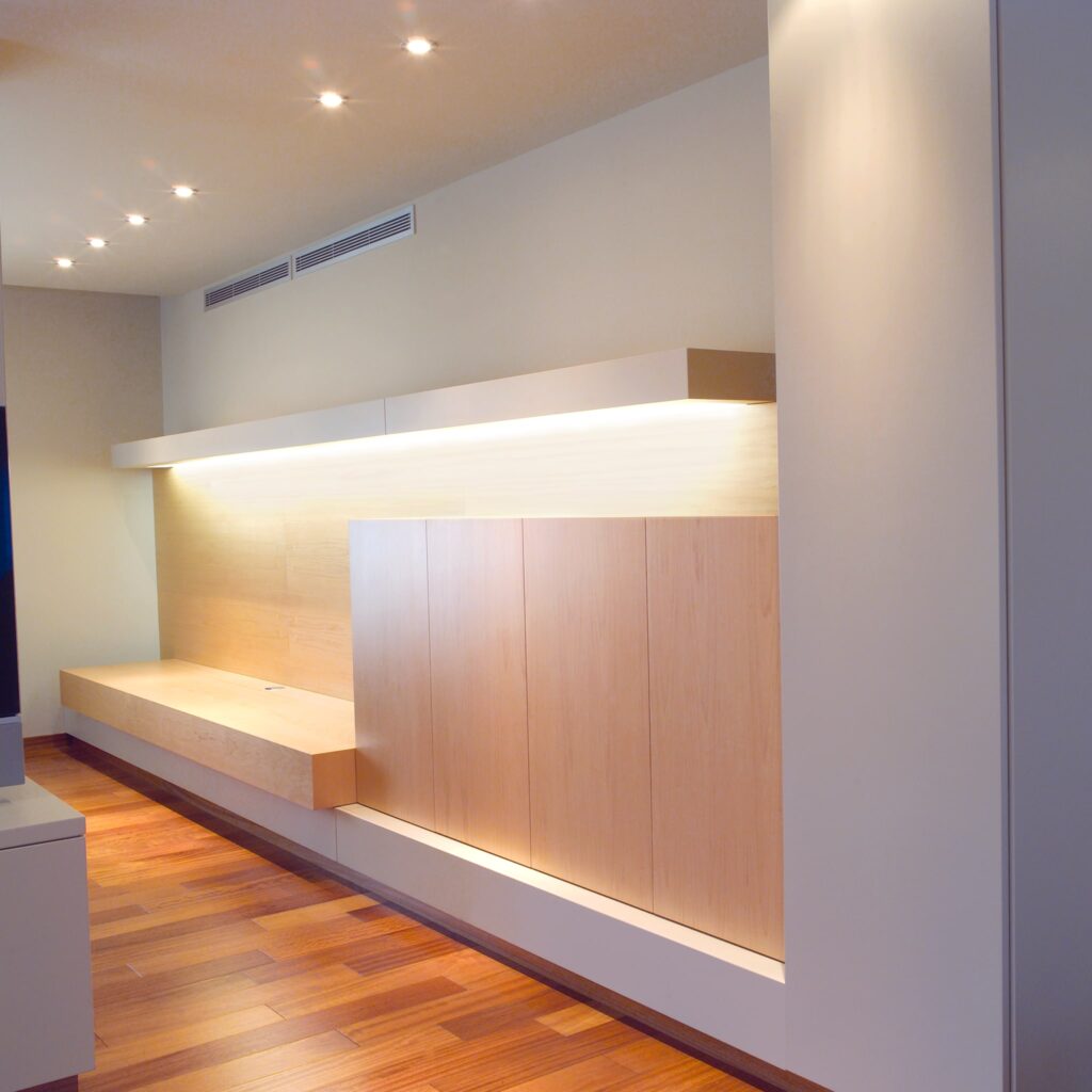 Diseño de interiores contemporáneo en Barcelona con mobiliario de alta gama Proyecto de diseño de interiores en Barcelona que muestra un banco de madera elegante con iluminación integrada, armarios minimalistas y un suelo de madera cálido, reflejando la esencia del diseño moderno y la arquitectura de alta gama.