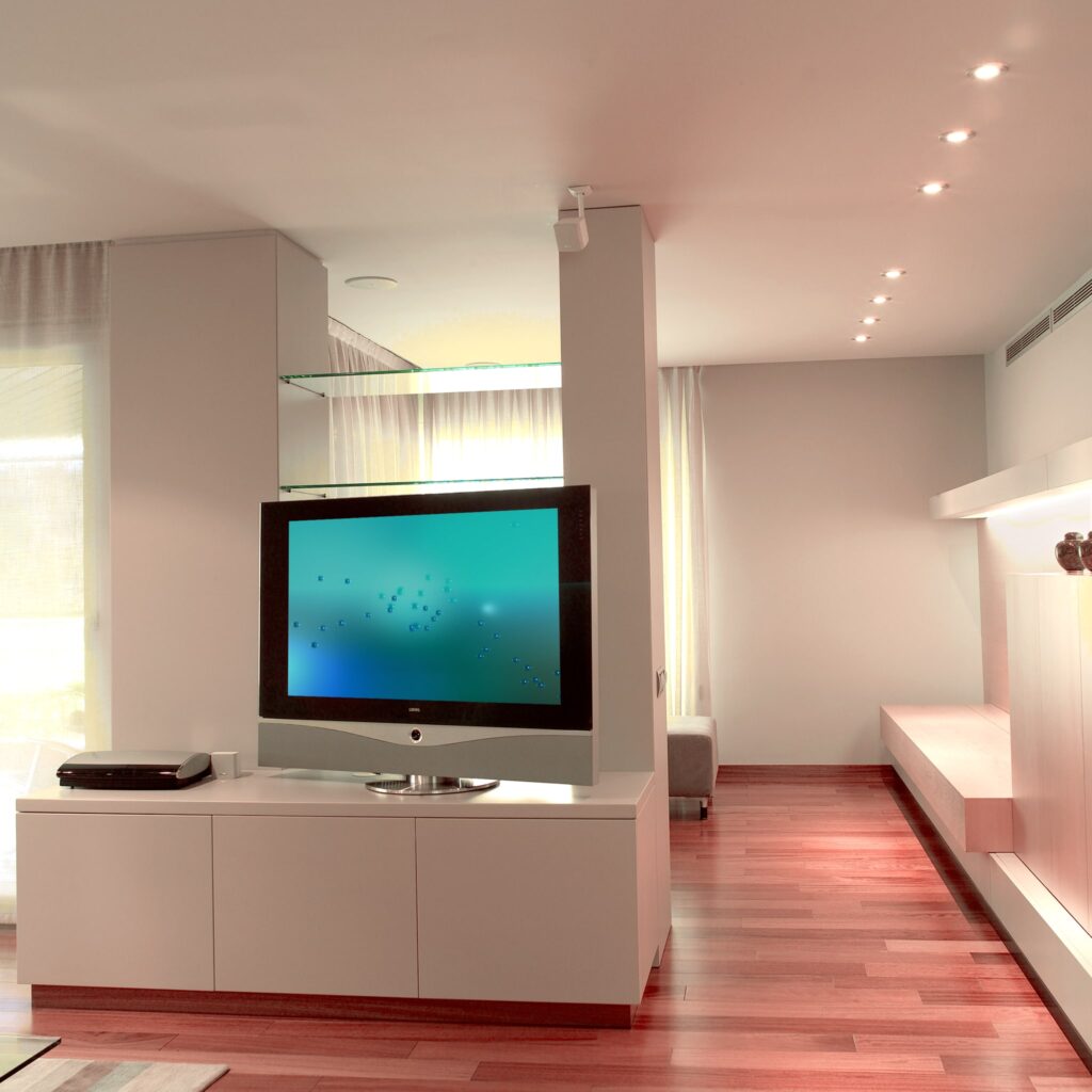 Sala de Estar Moderna con Televisor de Pantalla Plana y Luz Natural Sala de estar contemporánea que muestra un diseño elegante con un gran televisor de pantalla plana montado en un moderno mueble blanco brillante, bajo una suave luz empotrada cálida, con suelos de madera pulida y cortinas ligeras que permiten el paso de luz natural.