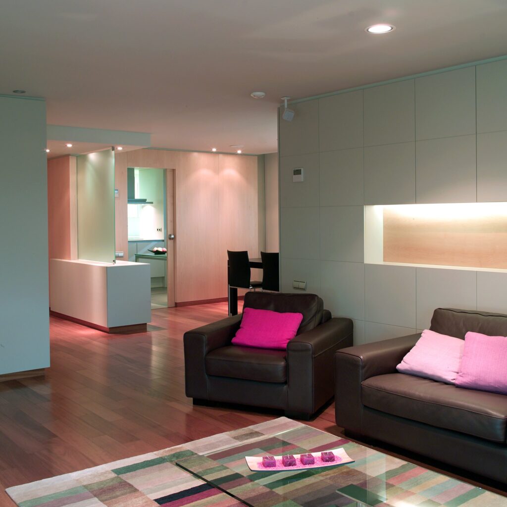 Espacio Habitacional Moderno con Sofás de Cuero Oscuro y Ambiente Acogedor Una sala de estar contemporánea con dos sofás de cuero oscuro y cojines rosas sobre una alfombra de área con un patrón, cálidamente iluminada por luces empotradas, con vista a una cocina minimalista en el fondo y un suelo de madera cálido.