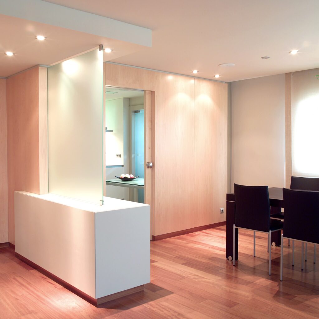 Interior de Espacio Abierto Contemporáneo con Suelo de Madera y Iluminación Cálida Una vista interior moderna que presenta un diseño espacioso con suelo de madera, una partición blanca minimalista con un panel de vidrio, una mesa de comedor con sillas negras y una encimera adornada con un cuenco de fruta, todo iluminado por una luz cálida.