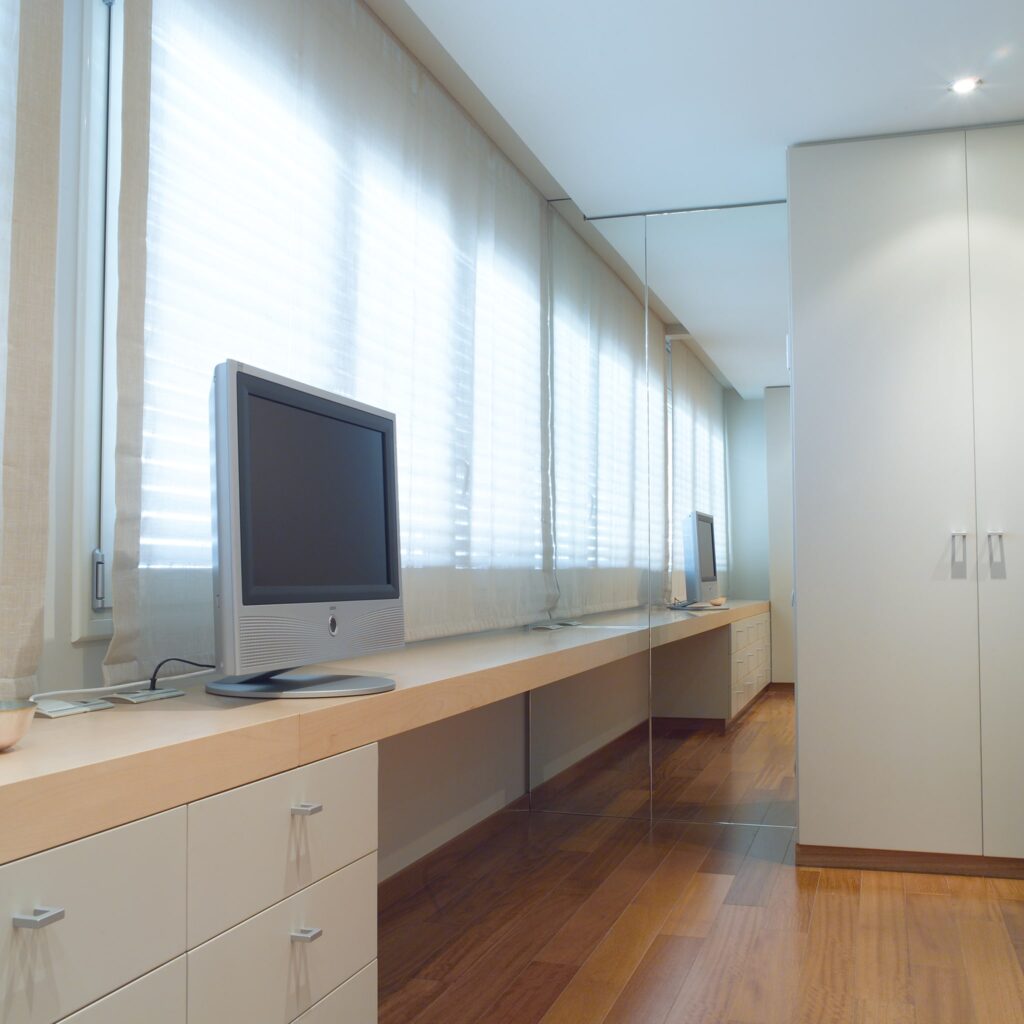 Diseño de Interiores de Oficina en Casa Moderno con Luz Natural Oficina en casa moderna, brillante y aireada, que presenta un elegante escritorio de madera con un televisor de pantalla plana, gabinetes claros y un cálido suelo de madera iluminado por la luz natural que entra a través de cortinas translúcidas.