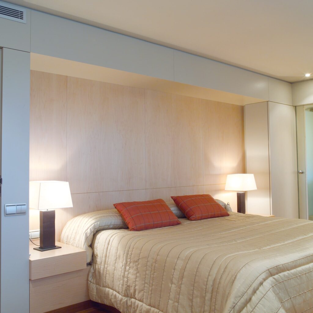 Dormitorio Moderno Acogedor con Edredón Beige Claro y Cojines de Cuadro Naranja Un dormitorio moderno y acogedor que presenta una cama bien hecha con un edredón beige claro y dos cojines de cuadro naranja, un cabecero integrado sin costuras con paneles de madera clara, mesas de noche pequeñas con lámparas brillantes y una iluminación cálida que crea una atmósfera serena.