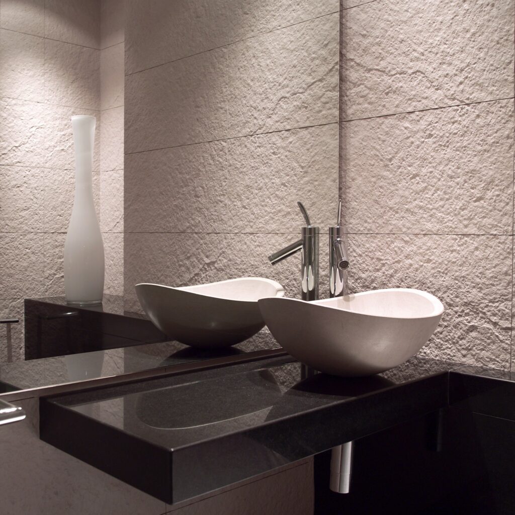 Baño Moderno Elegante con Lavabos de Tazón Blanco y Encimera Oscura Un baño moderno que presenta dos elegantes lavabos en forma de tazón blanco montados sobre una encimera oscura y estilizada, complementados por grifos de cromo y azulejos de pared de color claro texturizados. Un alto jarrón de vidrio esmerilado añade sofisticación al diseño minimalista.