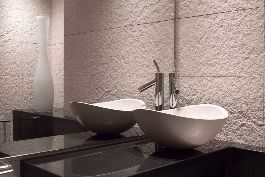 Un baño moderno que presenta dos elegantes lavabos en forma de tazón blanco montados sobre una encimera oscura y estilizada, complementados por grifos de cromo y azulejos de pared de color claro texturizados. Un alto jarrón de vidrio esmerilado añade sofisticación al diseño minimalista.