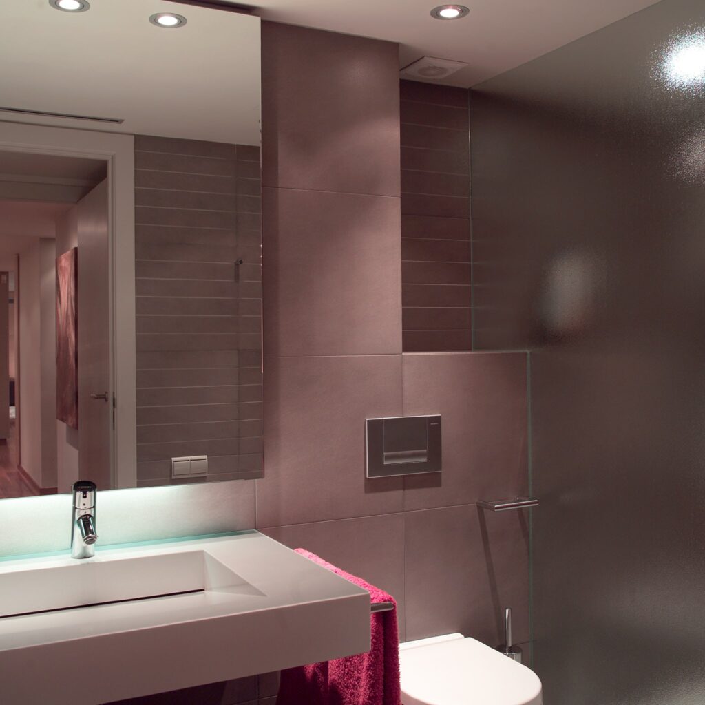 Diseño de baño moderno con lavabo minimalista y grifo elegante Un diseño de baño contemporáneo con un lavabo minimalista, grifo elegante y azulejos texturizados en suaves tonos neutros. Una toalla rosa cuelga cerca, y un gran espejo refleja la decoración estilosa, complementada por una suave iluminación empotrada. Un inodoro moderno es parcialmente visible a la derecha.