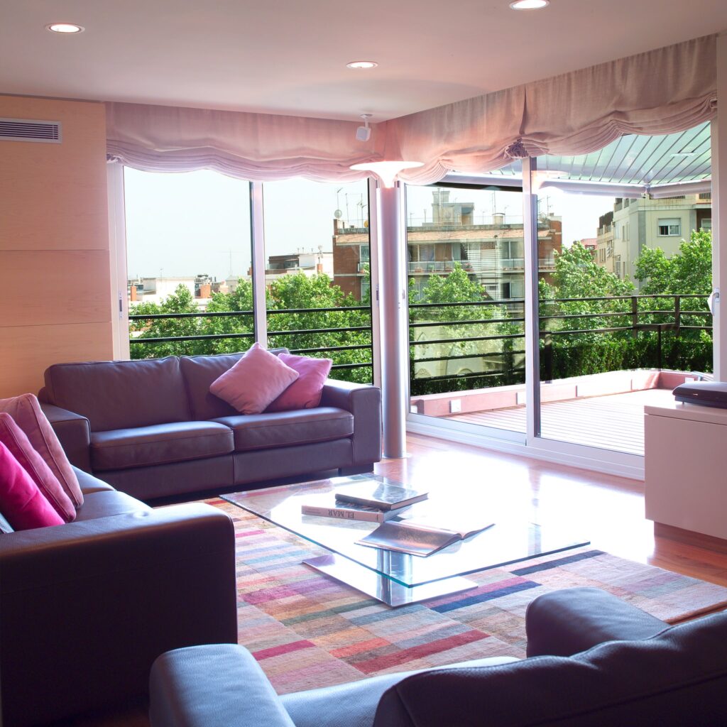 Piso en Calle de los Vergós: Amplia sala de estar moderna con grandes ventanas que ofrecen una vista de exuberante vegetación, dos sofás gris oscuro con cojines rosas, una mesa de café de vidrio y una colorida alfombra, iluminada por luz natural y acentos de madera.